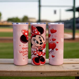 Op de afbeelding: Drie roze ge&iuml;soleerde bekers met zilveren deksels. E&eacute;n heeft Minnie Mouse met een hart, een andere heeft harten en de derde zegt "Happy Valentine's Day!" in het rood. De bekers staan op een houten oppervlak.