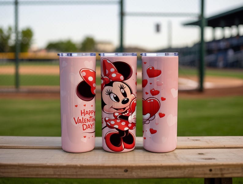 Op de afbeelding: Drie roze ge&iuml;soleerde bekers met zilveren deksels. E&eacute;n heeft Minnie Mouse met een hart, een andere heeft harten en de derde zegt "Happy Valentine's Day!" in het rood. De bekers staan op een houten oppervlak.