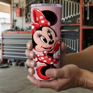 Op de afbeelding: Een roze beker met een Minnie Mouse-ontwerp. Minnie draagt een rode jurk met witte stippen en een strik. De cilindrische beker is waarschijnlijk gemaakt van roestvrij staal en is geschikt voor koude dranken.
