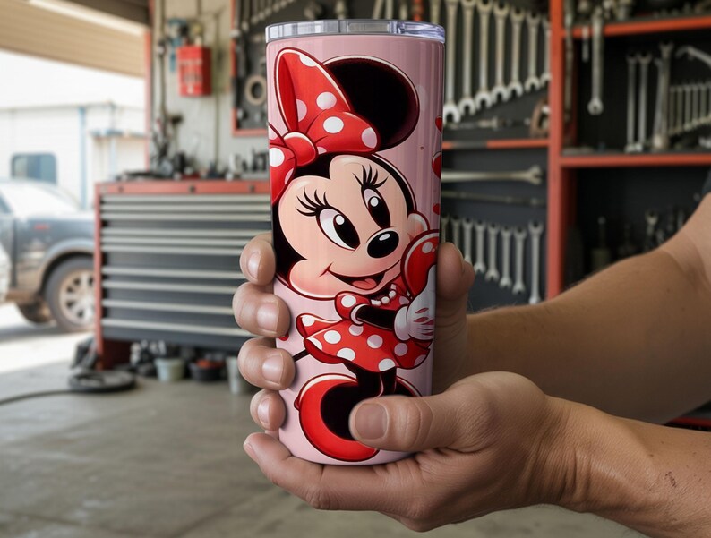Op de afbeelding: Een roze beker met een Minnie Mouse-ontwerp. Minnie draagt een rode jurk met witte stippen en een strik. De cilindrische beker is waarschijnlijk gemaakt van roestvrij staal en is geschikt voor koude dranken.