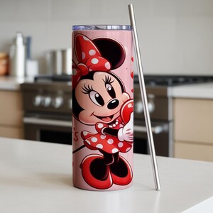 Op de afbeelding: Een roze beker met een Minnie Mouse-illustratie. Minnie draagt haar iconische rode jurk met witte stippen en een strik. Een roestvrijstalen rietje staat naast de beker. De beker is ontworpen voor koude dranken.