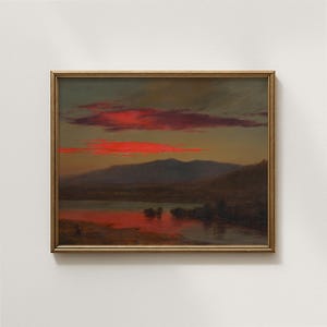 Op de afbeelding: Een ingelijst landschapsschilderij toont een zonsondergang boven een meer en bergen. De lucht is gevuld met levendige rode wolken, die weerspiegelen in het water. Het kunstwerk heeft een gouden lijst en een vintage esthetiek.