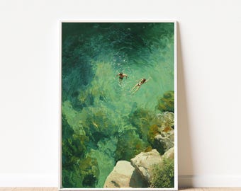 May include: A framed art print depicts two people swimming in clear turquoise water. The artwork showcases a high-angle view of the swimmers near a rocky coastline. The water's surface reflects sunlight, creating a shimmering effect. The print is displayed on a white wall above a wooden floor.