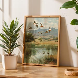 May include: A framed landscape painting depicts ducks in flight over a pond, with a backdrop of rolling hills and a cloudy sky. The artwork is in a wooden frame, displayed on a wooden table next to a potted plant.
