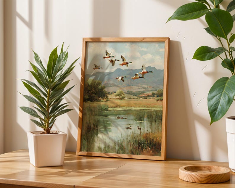 May include: A framed landscape painting depicts ducks in flight over a pond, with a backdrop of rolling hills and a cloudy sky. The artwork is in a wooden frame, displayed on a wooden table next to a potted plant.