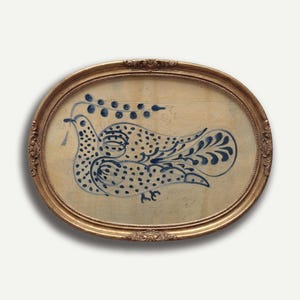 Puede incluir: Obra de arte enmarcada ovalada con una ilustración de paloma de arte popular azul y blanca. La paloma está adornada con lunares y detalles florales, sosteniendo una ramita en su pico. La obra de arte está enmarcada en un marco dorado ornamentado.