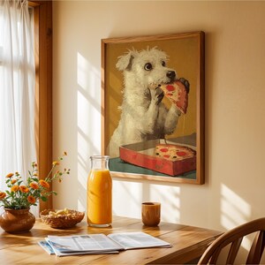 May include: A framed print shows a white dog eating pizza from a red box. The artwork hangs on a beige wall above a wooden table. A glass bottle of orange juice, a small brown cup, and flowers are on the table.