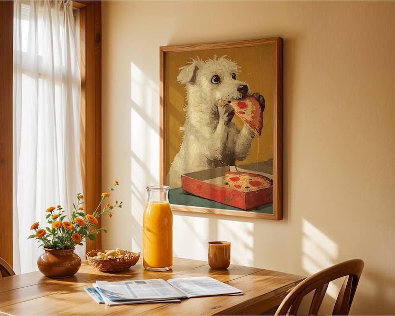 May include: A framed print shows a white dog eating pizza from a red box. The artwork hangs on a beige wall above a wooden table. A glass bottle of orange juice, a small brown cup, and flowers are on the table.