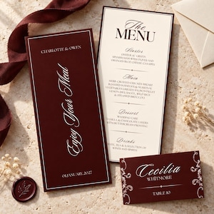 May include: A deep burgundy menu card with white text reading Charlotte & Owen, Enjoy Your Meal, and 01 January 2027, alongside a cream-colored menu listing courses and a burgundy place card for Cecilia Whitmore at Table 10 with white filigree.