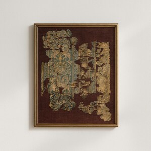 May include: Framed artwork featuring a fragment of antique textile. The textile, with faded teal and gold patterns, is mounted on a dark red background within a gold-toned frame. The textile shows signs of age and wear.