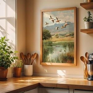 May include: A framed landscape painting depicts ducks in flight over a pond, with a wooden frame. The artwork is displayed in a kitchen setting, alongside potted plants, wooden utensils, and a coffee maker. The color palette includes greens, browns, and blues.
