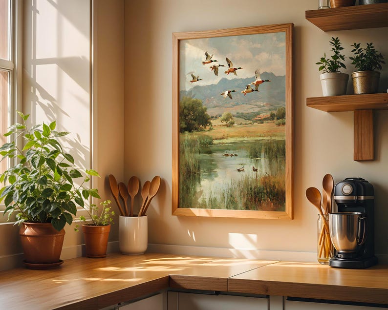 May include: A framed landscape painting depicts ducks in flight over a pond, with a wooden frame. The artwork is displayed in a kitchen setting, alongside potted plants, wooden utensils, and a coffee maker. The color palette includes greens, browns, and blues.