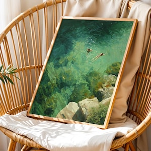 May include: A framed art print depicting two figures swimming in clear, turquoise water. The artwork, with a wooden frame, is displayed on a wicker chair with a white cushion. The scene captures a serene underwater view with visible rocks and sunlight.