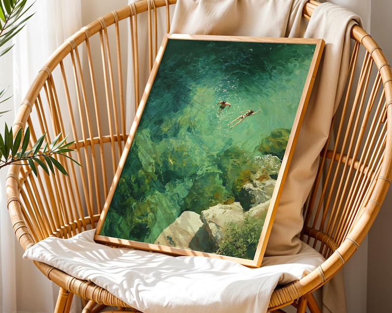 May include: A framed art print depicting two figures swimming in clear, turquoise water. The artwork, with a wooden frame, is displayed on a wicker chair with a white cushion. The scene captures a serene underwater view with visible rocks and sunlight.