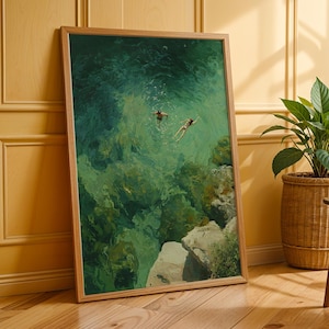 May include: A framed art print depicts two figures swimming in turquoise water near a rocky coastline. The artwork, with a wooden frame, is leaning against a yellow wall. A potted plant in a woven basket sits nearby.