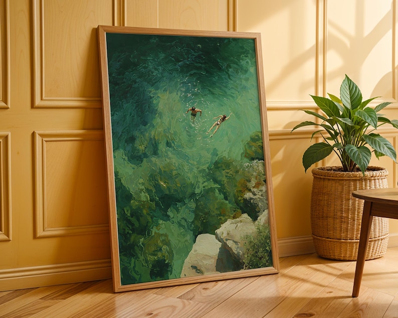 May include: A framed art print depicts two figures swimming in turquoise water near a rocky coastline. The artwork, with a wooden frame, is leaning against a yellow wall. A potted plant in a woven basket sits nearby.