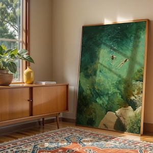 May include: A large framed art print depicts two figures swimming in turquoise water. The artwork leans against a wall next to a wooden cabinet with a potted plant and a yellow vase. A patterned rug lies on the wooden floor.