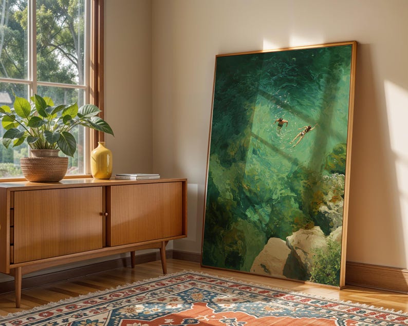 May include: A large framed art print depicts two figures swimming in turquoise water. The artwork leans against a wall next to a wooden cabinet with a potted plant and a yellow vase. A patterned rug lies on the wooden floor.