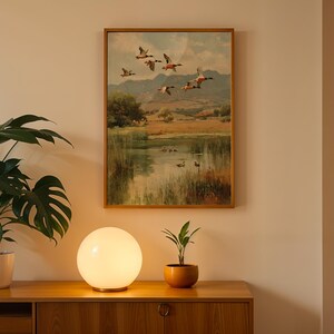 May include: A framed landscape painting depicts ducks in flight over a pond, with a wooden cabinet, a spherical lamp, and a potted plant in the foreground. The painting has a warm color palette, with a gold frame. The lamp emits a soft, warm glow.