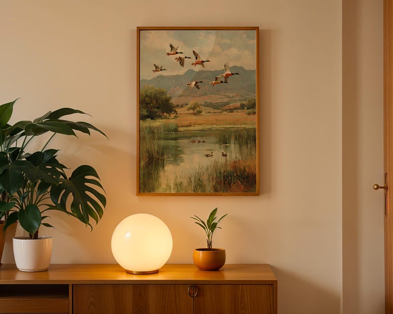 May include: A framed landscape painting depicts ducks in flight over a pond, with a wooden cabinet, a spherical lamp, and a potted plant in the foreground. The painting has a warm color palette, with a gold frame. The lamp emits a soft, warm glow.