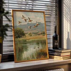 May include: A framed landscape painting featuring a flock of ducks in flight over a marshy lake. The artwork has a gold frame and depicts a serene natural scene with trees, mountains, and a body of water. A bottle of wine and books are next to the painting.