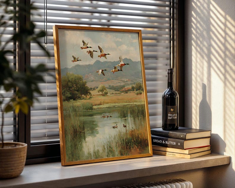 May include: A framed landscape painting featuring a flock of ducks in flight over a marshy lake. The artwork has a gold frame and depicts a serene natural scene with trees, mountains, and a body of water. A bottle of wine and books are next to the painting.
