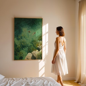 May include: A framed painting with a gold border depicts two figures swimming in turquoise water. A woman in a white dress stands in front of the artwork, near a bed with white linens. Sunlight streams through a window.