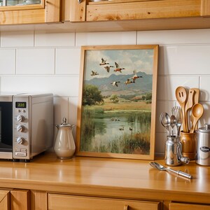 May include: A framed landscape painting of ducks in flight over a pond, with a wooden frame. The painting is displayed on a wooden countertop next to a stainless steel microwave, a creamer, and kitchen utensils.