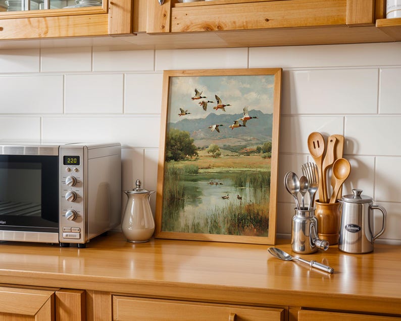 May include: A framed landscape painting of ducks in flight over a pond, with a wooden frame. The painting is displayed on a wooden countertop next to a stainless steel microwave, a creamer, and kitchen utensils.