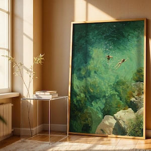 May include: A framed art print depicts two figures swimming in turquoise water. The artwork, with a gold frame, is displayed against a beige wall. The scene captures a serene moment with sunlight streaming through a nearby window.