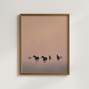 May include: Framed art print featuring a silhouette of several horses running across a misty landscape. The artwork has a warm, muted color palette with a gold-colored frame. The horses are dark against a soft, peachy-pink sky.