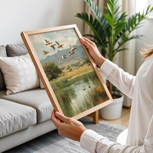 May include: A framed landscape painting featuring a flock of ducks in flight over a serene lake and rolling hills. The artwork is set in a light wooden frame, showcasing a classic, nature-themed scene with muted colors.