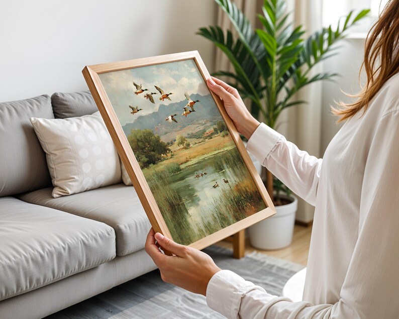 May include: A framed landscape painting featuring a flock of ducks in flight over a serene lake and rolling hills. The artwork is set in a light wooden frame, showcasing a classic, nature-themed scene with muted colors.