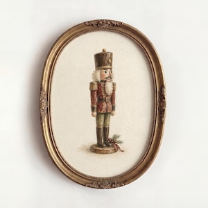 May include: An oval-shaped, ornate gold frame containing a detailed illustration of a nutcracker soldier. The soldier wears a brown hat, a red jacket with gold epaulets, and dark trousers, standing on a circular base with a small pine sprig.