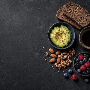 Könnte beinhalten: Ein Frühstücksarrangement mit zwei Scheiben dunklem Roggenbrot, einer kleinen Schüssel mit in Scheiben geschnittener Avocado und schwarzem Pfeffer, einer Tasse schwarzem Kaffee, einer Handvoll Mandeln und Walnüssen sowie frischen Beeren in einer Schale.