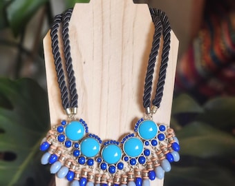 May include: A statement necklace with a black rope chain and a bib pendant. The pendant features large turquoise blue cabochons, smaller blue beads, and gray teardrop-shaped beads. The necklace is displayed on a wooden jewelry stand.