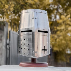 May include: A silver-colored, riveted knight's helmet with a cross emblem, mounted on a wooden stand. The helmet features eye slits and ventilation holes. The text "VALORFORGEIN" is visible on the side.