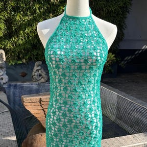 Puede incluir: Vestido de ganchillo verde con cuello halter, exhibido en un maniquí. El vestido tiene una silueta ajustada y un delicado patrón de tejido abierto. El vestido mide aproximadamente 91 cm de largo.