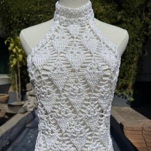 Pode incluir: Um top de crochê branco estilo halter. O top apresenta uma gola alta e um padrão de trabalho aberto detalhado. A peça de roupa é exibida em um manequim, mostrando sua textura e design. O fundo inclui vegetação e um elemento aquático.