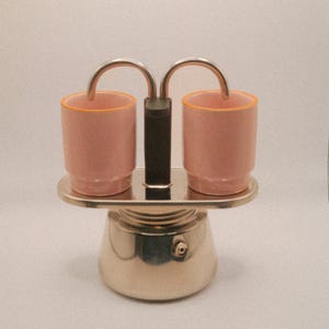 May include: A silver stovetop espresso maker with two pink cups. The cups have orange rims and are connected by a silver handle. The base of the coffee maker is silver and has a black handle.