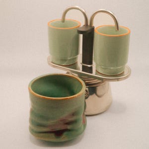 May include: A stainless steel stovetop espresso maker with two small green cups with orange rims on top. A larger green ceramic cup with a textured surface sits in front. The cups and espresso maker are on a white surface.