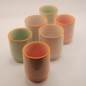 May include: Six ceramic cups in pastel colors: light green, pink, and peach. Each cup has a contrasting orange rim. The cups are arranged in a slightly offset pattern, showcasing their design and color variations.