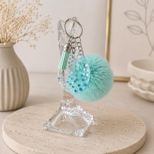 May include: A keyring featuring a silver metal ring, a light mint green tassel, a clear circular acrylic charm with blue and mint green polka dot patterns, and a fluffy, round, light mint green faux fur pom-pom attached to the chain.