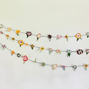 Op de afbeelding: Een kleurrijke bloemenkrans met rode, gele, oranje, witte en groene bloemen. De krans is opgehangen aan een witte muur met een witte deur op de achtergrond.