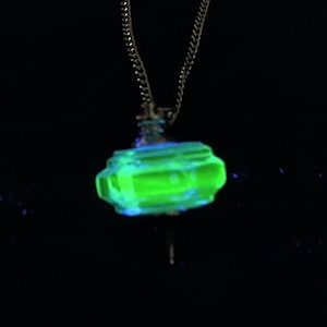 Puede incluir: Collar con colgante verde brillante y cadena plateada. El colgante tiene una forma inusual e irregular con detalles azules, sobre un fondo negro. El colgante parece estar hecho de un material luminoso.
