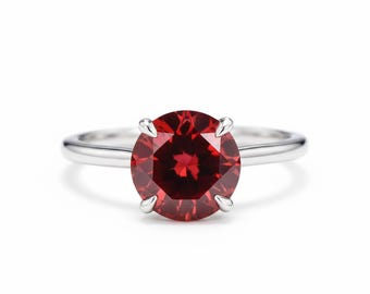 Puede incluir: Un anillo de plata con una gran gema roja oscura, redonda y facetada. La gema está sujeta por cuatro garras. La banda del anillo es lisa y pulida, reflejando la luz. Diseño clásico.