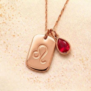 以下が含まれることがあります： 長方形のペンダントに獅子座のシンボルと涙型の赤い宝石が付いたローズゴールドのネックレス。ペンダントと宝石は繊細なチェーンから吊り下げられており、エレガントでパーソナライズされたアクセサリーとなっています。