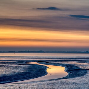 Op de afbeelding: Een landschapsfoto van een kustgebied bij zonsondergang. De lucht gaat over van oranje naar paars, weerspiegeld in het water. Een kronkelend waterkanaal snijdt door het zand en leidt naar de horizon.