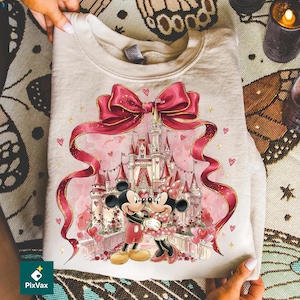 May include: A beige sweatshirt featuring a romantic design with Mickey and Minnie Mouse in front of a castle. The design includes a large pink bow, ribbons, hearts, and gold accents. The couple is embracing, with a castle in the background.