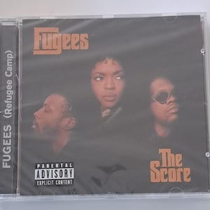 May include: A CD case featuring the album "The Score" by the Fugees. The cover art displays three band members against a dark background, with the band's name and album title in orange. A parental advisory warning is visible.